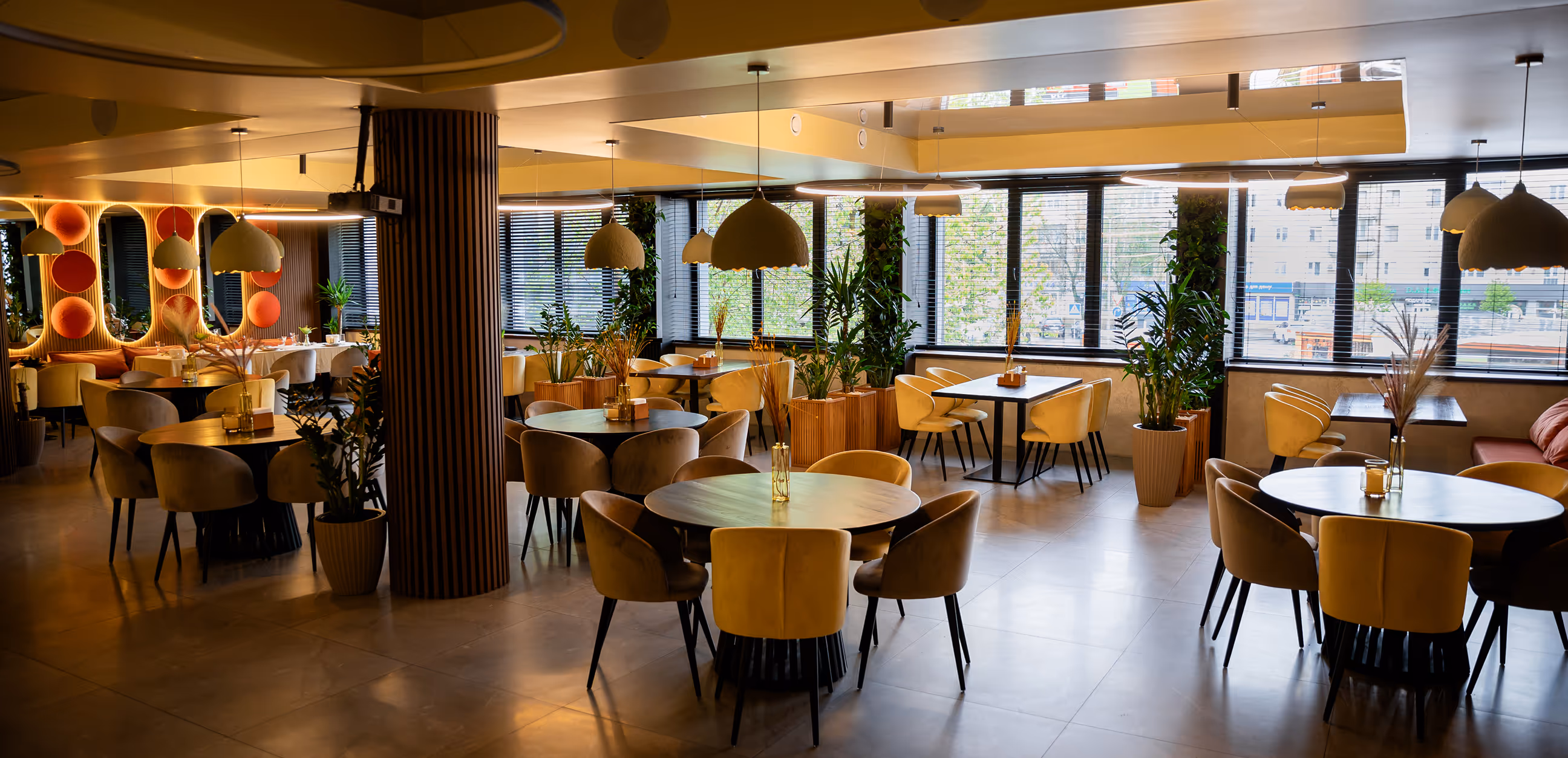 Modern restaurant interior with round and square tables surrounded by beige and mustard chairs, pendant lighting, and large windows with plants.
