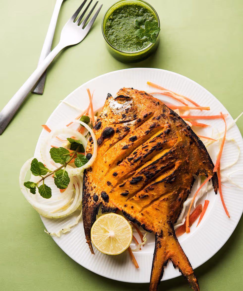 Grilled pomfret fish with lemon half, onion rings, carrot strips, and mint leaves on a white plate, served with green chutney in a glass.
